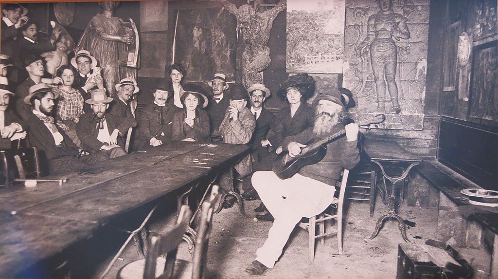 Frederic Gerard known as Frede the landlord of the Lapin Agile cabaret in Montmartre plays the guitar on front of an attentive audience gathered around a table. The room is sparsly decorated with wooden chairs and tables. In the background Picasso’s painting of a harlequin and companinion known as At the Lapin Agile can be seen on the wall.