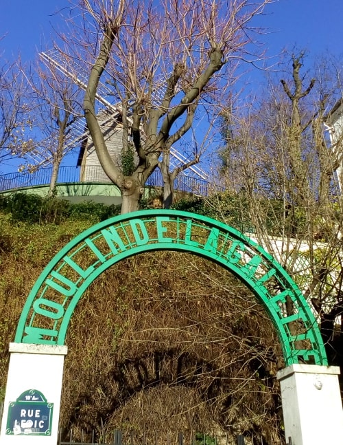The Blute-Fin windmill stands on the top of Montmartre in some gardens. The windmill is set behind some trees and is framed through an arched green sign announcing the Moulin de la Galette. The view is from below in Rue Lepic 75018 Paris.