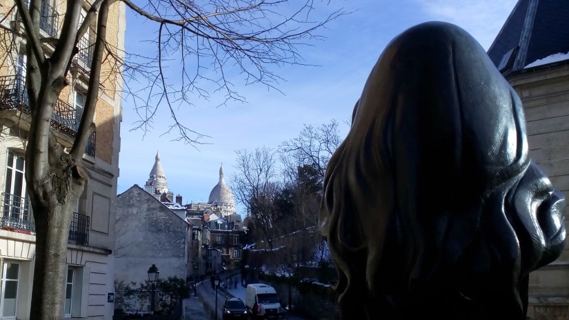 The bronze bust of French singer Dalida looks towards the Sacre Coeur Montmartre. The view is from behind her head. In the distance we see the dome of the Sacré Coeur Paris.