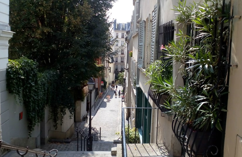 Looking down Rue André Antoine from Rue des Abbesses Montmartre. A series of stairs leads down to a narrow street fringed by shuttered buildings and plants. Railings and a traditional gas type street light can be seen in foreground.