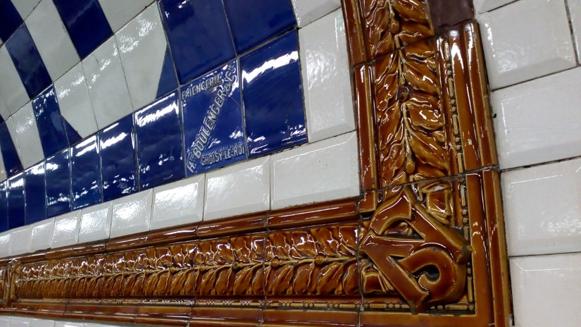 The detailed ceramic work around the station names on the old North South métro line. A brown ceramic tile bears the initials NS. Further ceramics surround the station name which is picked out in blue tiles on a white background. The North South line dates from 1911 and is now known as line number 12.