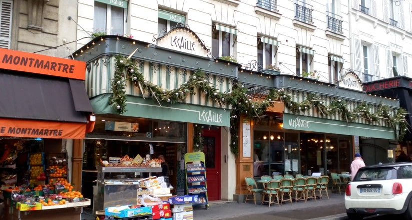 Rue des Abbesses is a lively street in the heart of Montmartre. It features many cafés and small businesses. The view shows a restaurant and a corner shop with fruit piled at the entrance.