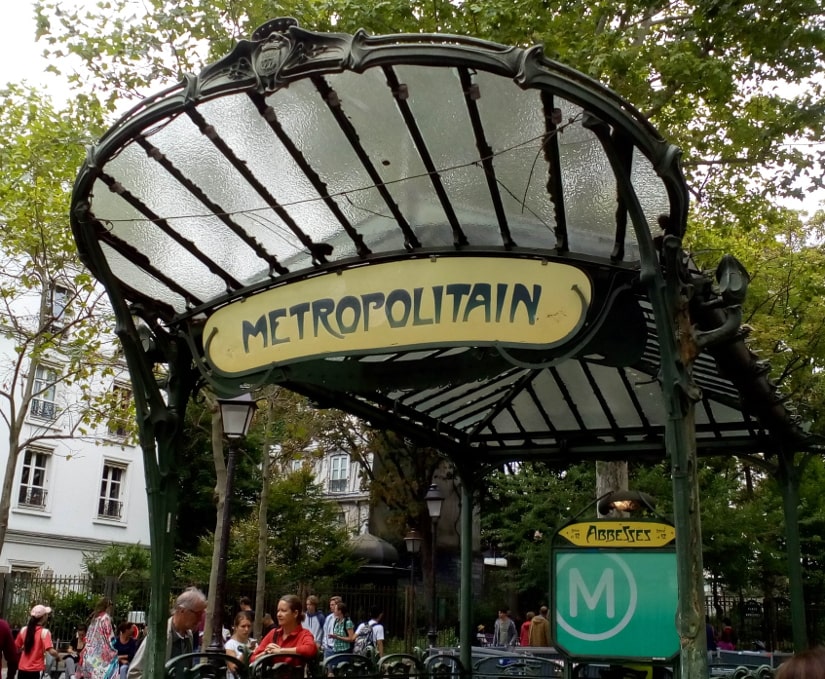 The Art Nouveau entrance to the Abbesses metro in Montmartre Paris. A glass and wrought iron structure shelters the steps leading down to the metro. The green structure is set in a busy wooded square with many tourists around the entrance.