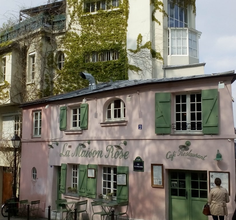 A small pink restaurant known as the Pink House in Montmartre. The windows have green shutters and some tables are set on the pavement. Adjoining the building and set behind are some taller ivy clad apartment blocks with balconies and large artist studio style windows.