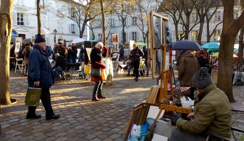 Place du Tertre Montmartre in winter. A painter sits with his paintings displayed on an easel. More painters can be seen behind sitting below umbrellas. In the middle ground tree trunks can be seen throughout the square and two tourists wander by the artist’s stalls.