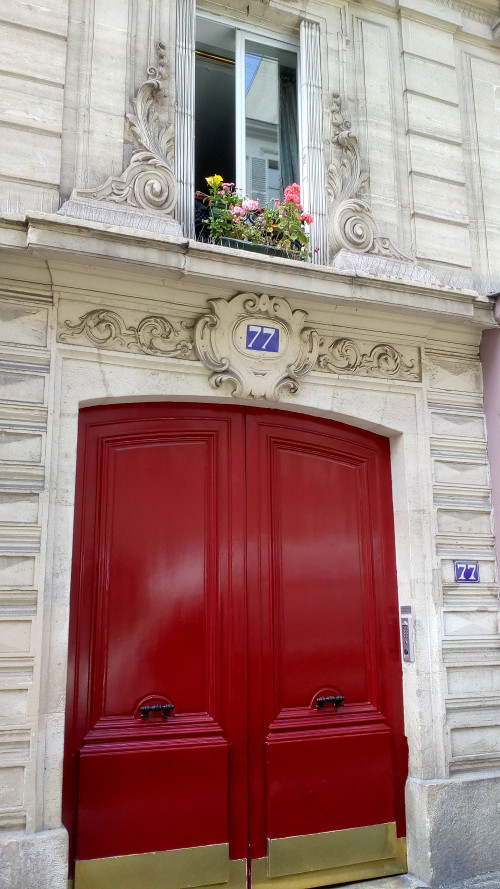 The bright red brass plated doors of 77 Rue Blanche Montmartre. This is one of Degas’ many apartments and studios in Montmartre. The number 77 is prominently displayed on a blue background and a window is framed in some florid decorative carving. A flower box is visible on the window ledge.