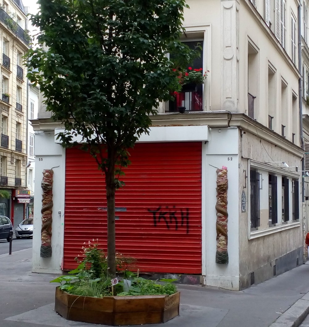 A closed shuttered red door with Chinese style decorations around it on front of which is a tree set in a small square. The building is the ground floor of an apartment block which forms the angle of Rue Jean-Baptiste Pigalle and Rue de la Rochfoucauld Montmartre. This is the site of le Grand Duc jazz night club in the 1920s
