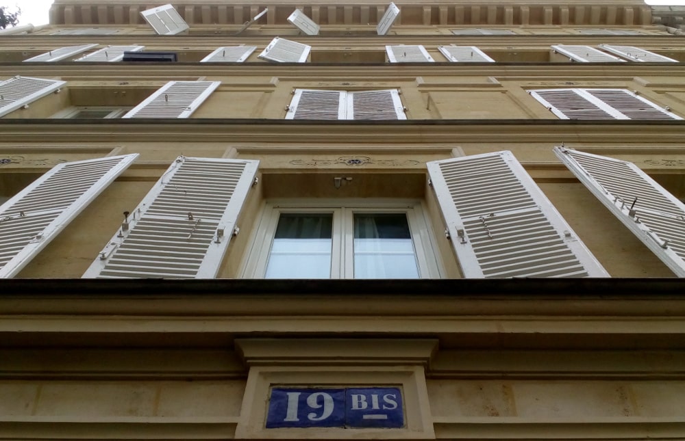 Looking up the facade of 19 Bis Rue Pierre Fontaine Montmartre. Windows and shutters are visible on the four storey building. Some shutters are thrown open. At the top is a cornice. The building is made from sandy yellow coloured stone.