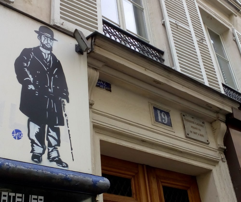 A stencilled image of Toulouse-Lautrec and cane stands next to the top of the door of his apartment at 19 Rue Pierre Fontaine Montmartre. Window shutters, the street number 19 and a plaque which tells us that Toulouse-Lautrec lived here are also visible.