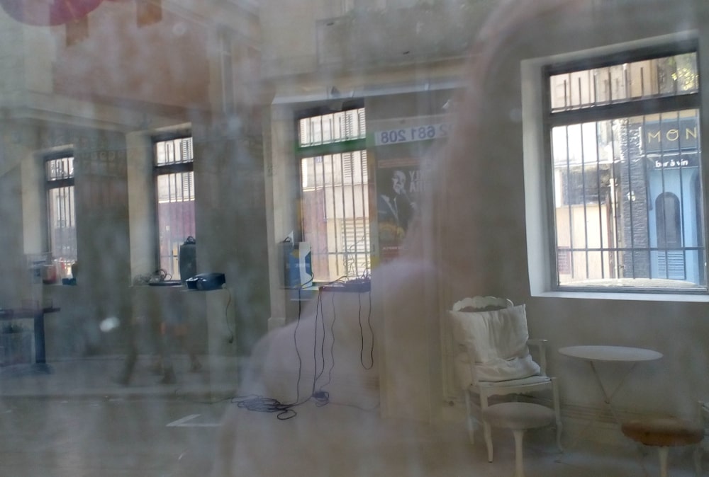 Looking through some windows at 52 Rue Jean-Baptiste Pigalle to the interior of what used to be the Grand Duc (Nightowl) jazz club in 1920s Jazz Age Paris. The interior is white with four barred windows. A chair a table and some electrical equipment is visible.