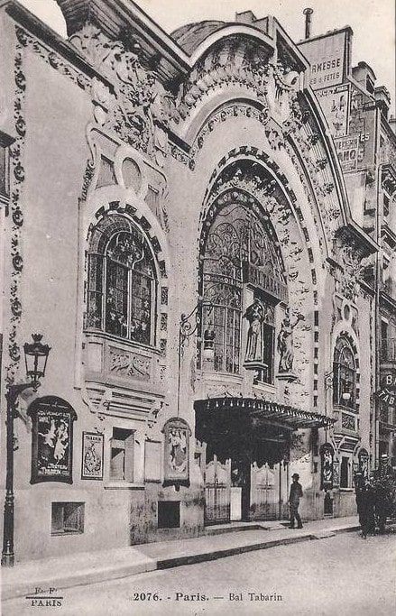 A huge arched window dominates the entrance to the Bal Tabarin. The building featured Art Nouveau sculpture and relief on the facade including two large smiling faces. A horse and cart are seen in front of the building as a man in a bowler hat enters.