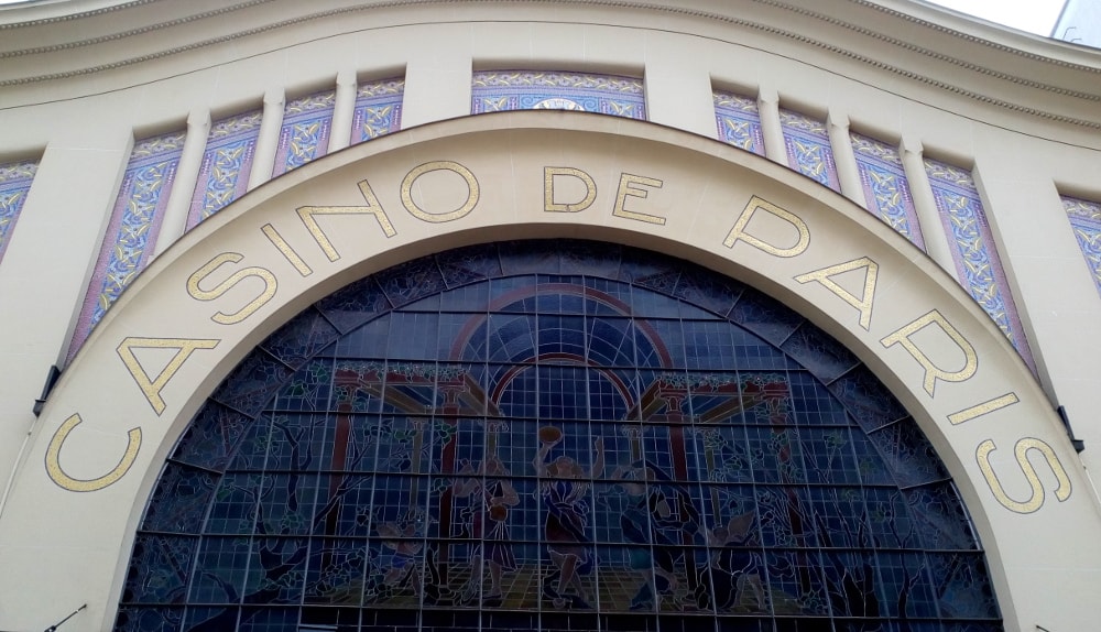 The white Art Deco stucco facade of the Casino de Paris concert hall. A large arched stained glass window dominates the facade. Casino de Paris is spelled out in large mosaic letters above the arch. Further blue mosaic decoration is visible above the arch. The arch movement is continued to the corniced roof giving a wave effect.