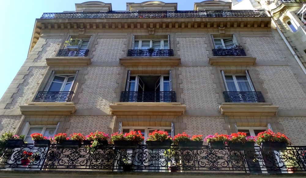 Degas 21 Rue Jean-Baptiste Pigalle. Looking up the facade of a brick and stone apartment building with a large balcony running across all of one floor. Flower boxes and flowers hang over the balcony. A large cornice is visible on the last floor on which runs another balcony.