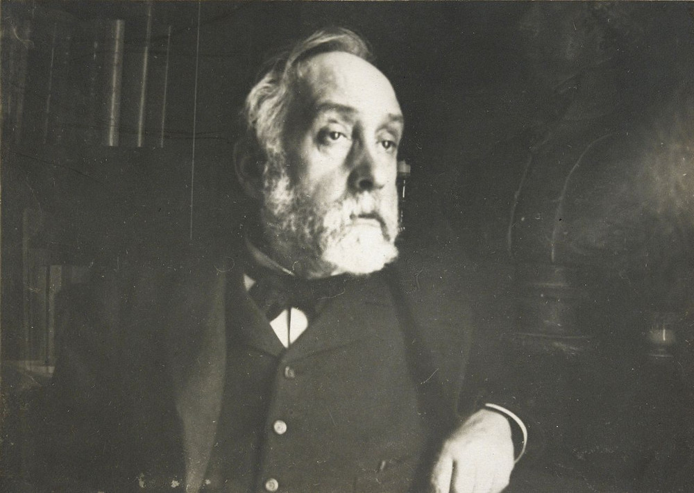 Degas’ photographic self portrait from 1895. A bearded and solemn looking man looks to the viewers right. He is wearing a jacket, waistcoat and cravat. Some sombre details of furniture are just visible in background.