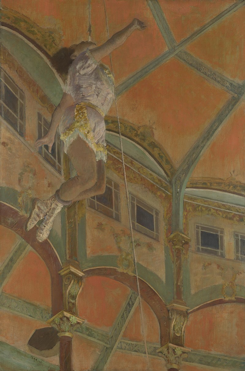 The circus performer Miss La La is dangling by her teeth above the spectators from near the roof of the Circus Fernando. Miss La La’s hands and feet are not attached to the rope from which she hangs. The ornate circus roof is visible behind her.